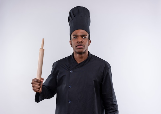 Young annoyed afro-american cook in chef uniform holds rolling pin isolated on white background with copy space