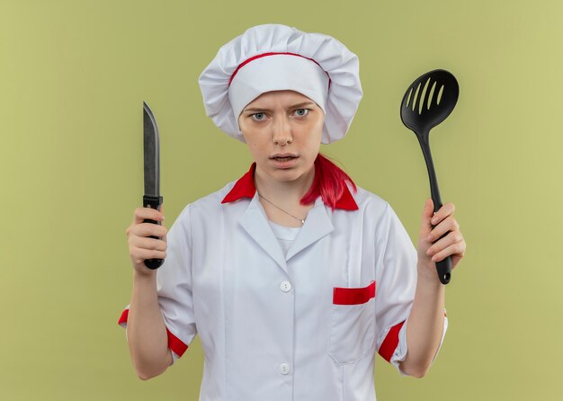 Young annoyed blonde female chef in chef uniform holds knife and spatula isolated on green wall