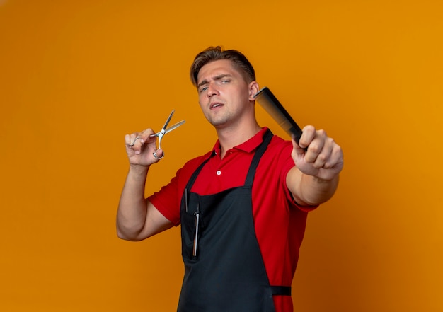 Young annoyed blonde male barber in uniform holds comb and scissors isolated on orange space with copy space