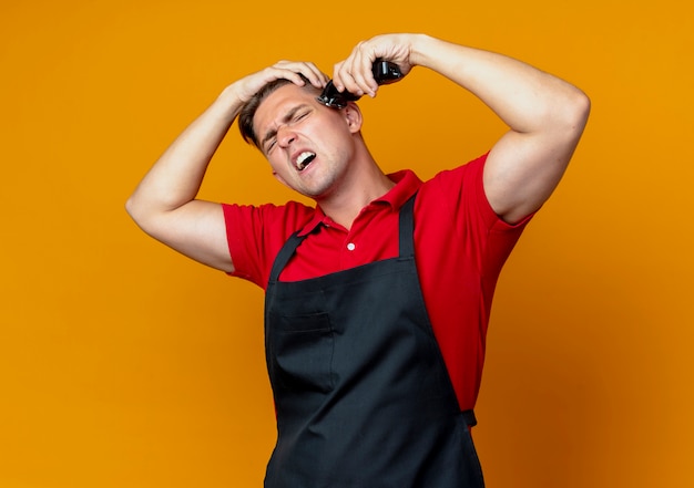 Free photo young annoyed blonde male barber in uniform holds head trimming hair with hair clipper isolated on orange space with copy space