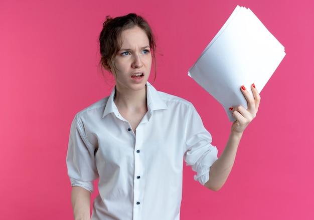 Young annoyed blonde russian girl holds paper sheets looking at side isolated on pink space with copy space