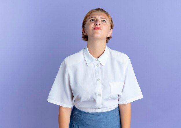 Young annoyed blonde russian girl looks up isolated on purple background with copy space