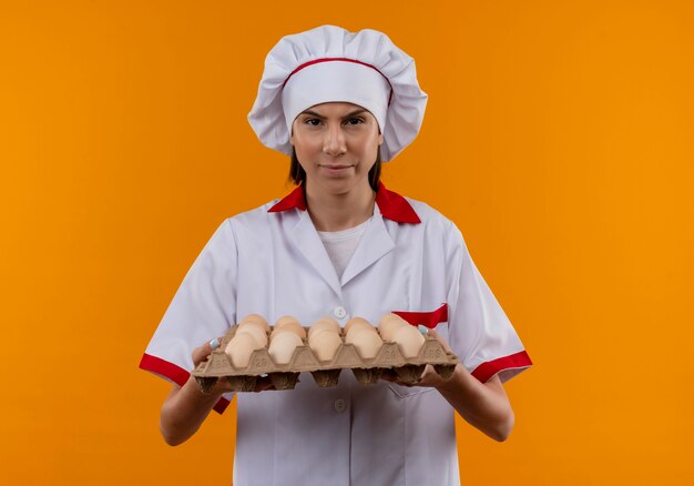 Young annoyed caucasian cook girl in chef uniform holds batch of eggs and looks at camera isolated on orange space with copy space