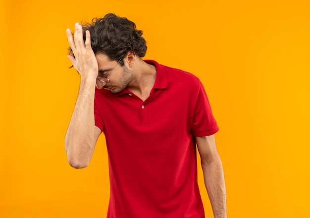 Young annoyed man in red shirt with optical glasses holds head and looks at side isolated on orange wall