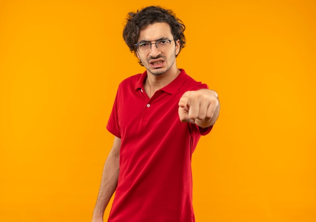 Young annoyed man in red shirt with optical glasses points isolated on orange wall