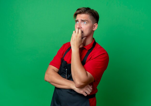 Young anxious blonde male barber in uniform bites fingers looking at side isolated on green space with copy space