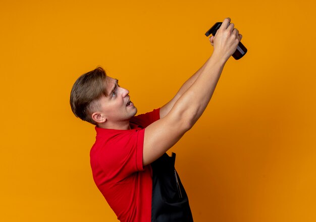 Young anxious blonde male barber in uniform stands sideways holding spray bottle isolated on orange space with copy space