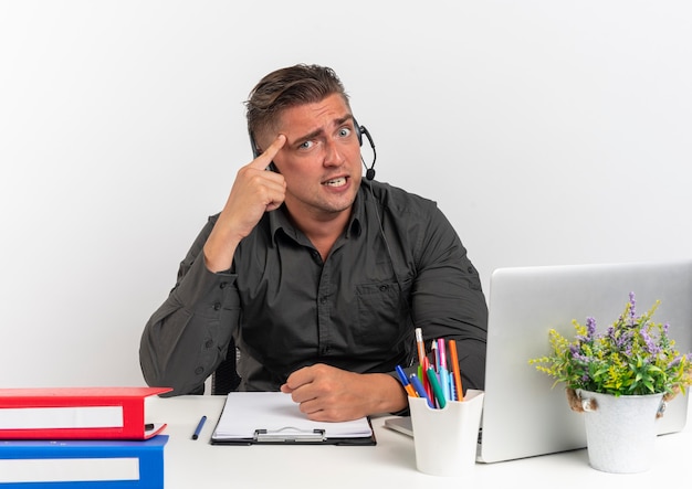 Young anxious blonde office worker man on headphones sits at desk with office tools using laptop puts finger on head looking at camera isolated on white background with copy space