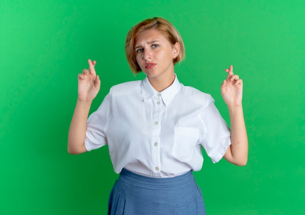 Free photo young anxious blonde russian girl crosses fingers isolated on green background with copy space