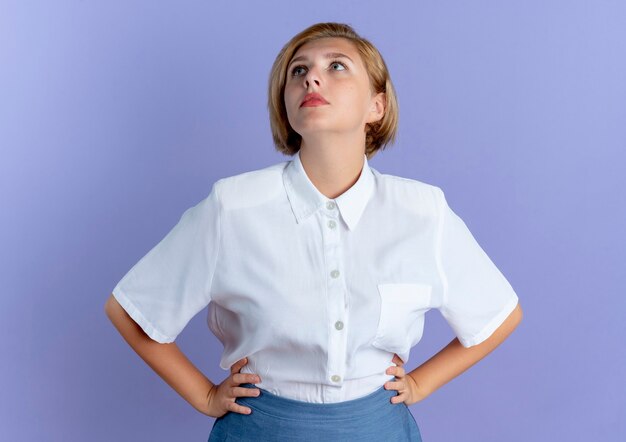 Young anxious blonde russian girl puts hands on waist looking up isolated on purple background with copy space