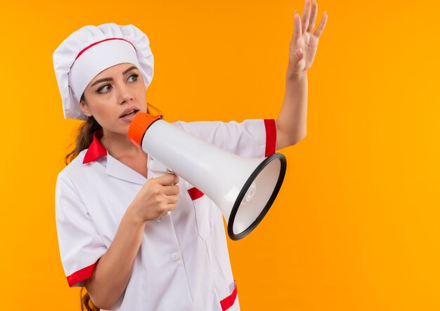 Young anxious caucasian cook girl in chef uniform holds loud speaker and raises hand up isolated on orange background with copy space