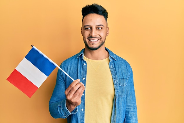 Free photo young arab man holding france flag looking positive and happy standing and smiling with a confident smile showing teeth