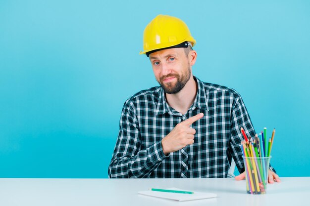 Young architect is looking at camera by pointing right with forefingers on blue background