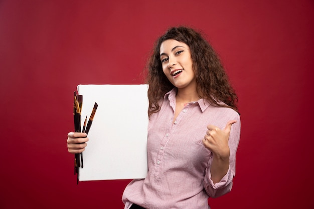 Young artist holding paintbrushes and canvas giving thumbs up.