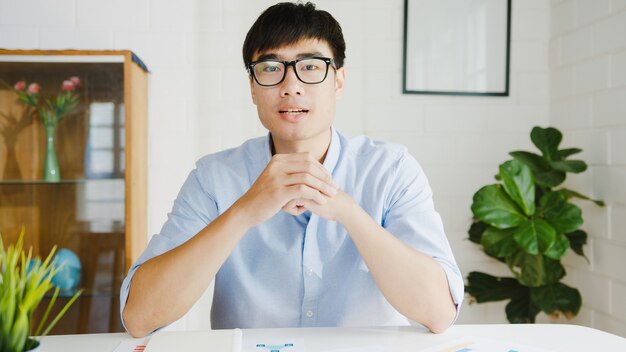 Young Asia businessman using computer laptop talk to colleagues about plan in video call meeting while working from home at living room. Self-isolation, social distancing, quarantine for corona virus.