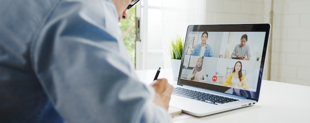 Young Asia businessman using laptop talk to colleagues about plan in video call meeting while work from home at living room. Self-isolation, social distancing, quarantine for corona virus prevention.