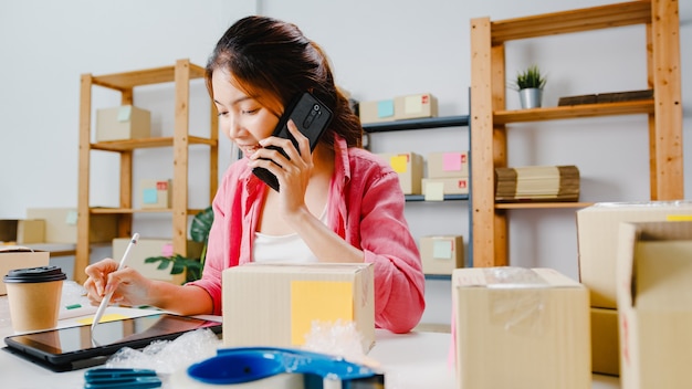 Young Asia businesswoman using mobile phone call receiving purchase order and check product on stock, work at home office. Small business owner, online market delivery, lifestyle freelance concept.