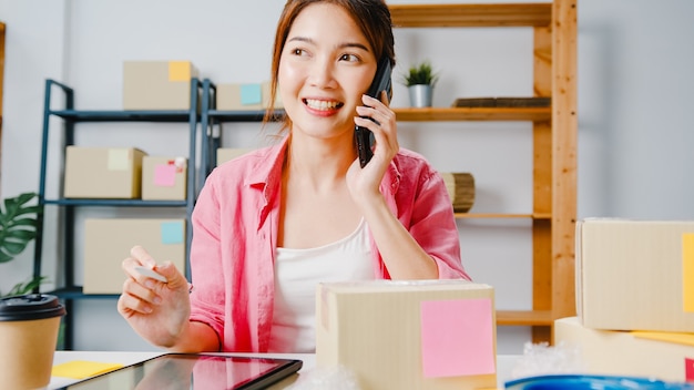 Young Asia businesswoman using mobile phone call receiving purchase order and check product on stock, work at home office. Small business owner, online market delivery, lifestyle freelance concept.