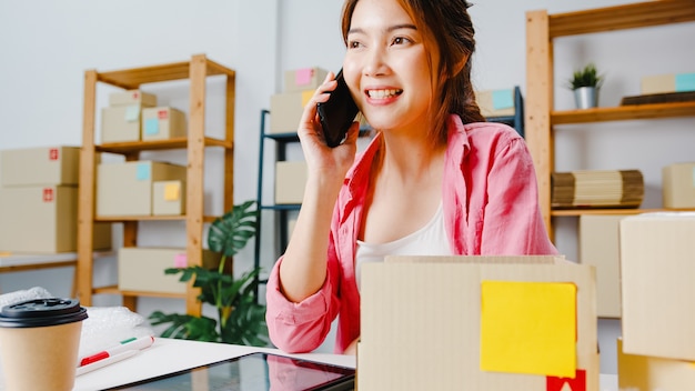 Young Asia businesswoman using mobile phone call receiving purchase order and check product on stock, work at home office. Small business owner, online market delivery, lifestyle freelance concept.