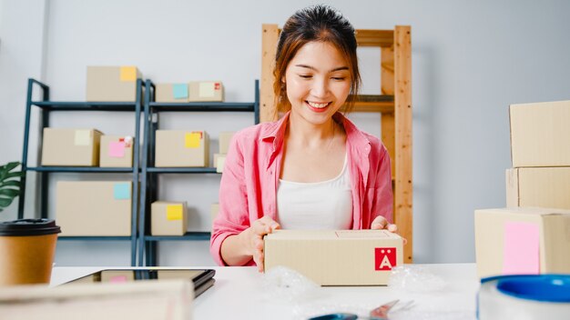 Young Asia entrepreneur businesswoman packing product in cardboard box deliver to customer, working at home office. Small business owner, start up online market delivery, lifestyle freelance concept.