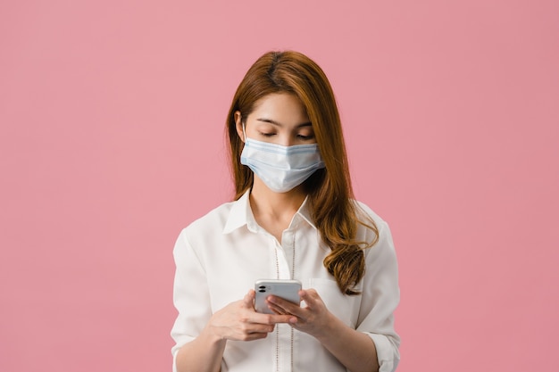 Young Asia girl wearing medical face mask using mobile phone with dressed in casual clothing isolated on pink background.