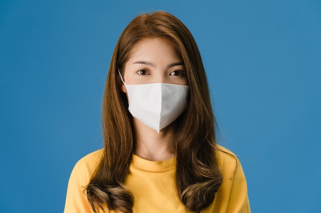 Young Asia girl wearing medical face mask with dressed in casual clothing and looking at camera isolated on blue background. Self-isolation, social distancing, quarantine for corona virus prevention.