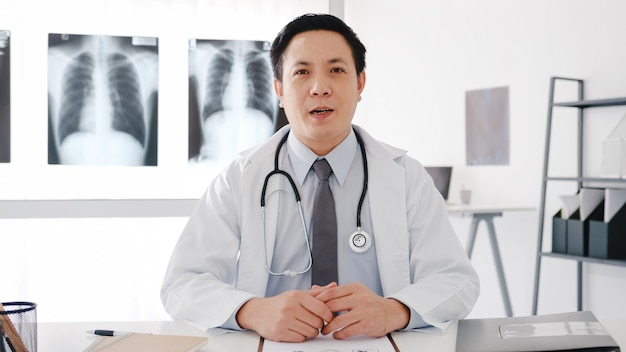 Young Asia male doctor in white medical uniform with stethoscope using computer laptop talk video conference call with patient, looking at camera in health hospital.