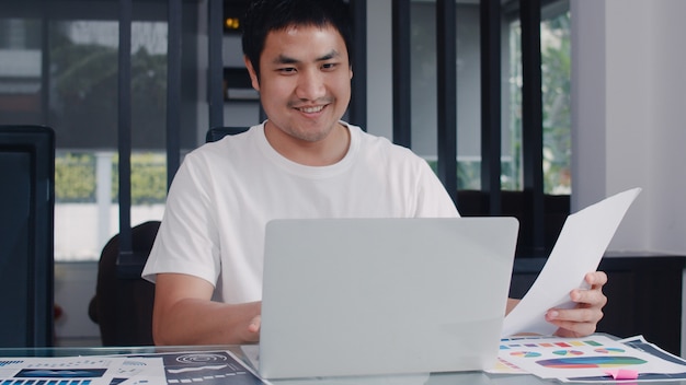 Young Asian business man records of income and expenses at home. male worried, serious, stress while using laptop record budget, tax, financial document working in living room at home .