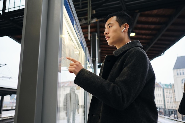 Young Asian businessman in wireless earphones thoughtfully watching timetable of public transport at metro station outdoor