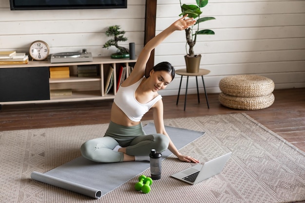 Young asian fitness girl doing stretching sport workout at home stretch arms and looking at laptop f...