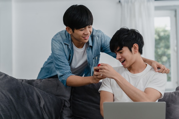 Young Asian gay couple propose at modern home, Teen korean LGBTQ men happy smiling have romantic time while proposing and marriage surprise wear wedding ring in living room at house .