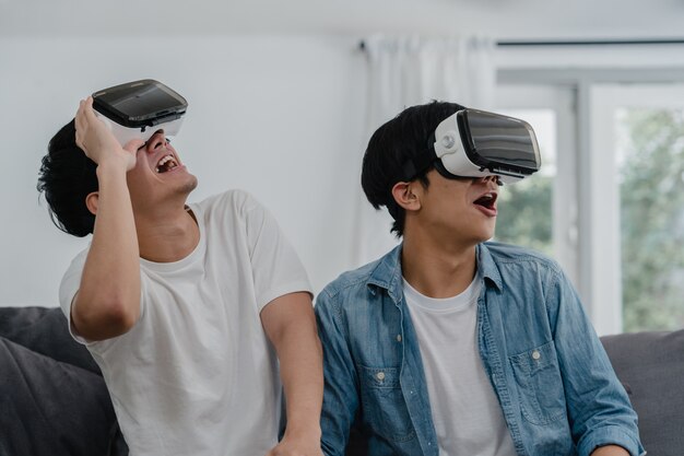 Young Asian Gay couple using technology funny at home, Asia lover guy lgbtq+ feeling happy fun and virtual reality, VR playing games together while lying sofa in living room at home .