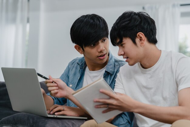 Young Asian Gay couple working laptop at modern home. Asia LGBTQ+ men happy relax fun using computer and analyzing their finances in internet together while lying sofa in living room at house .