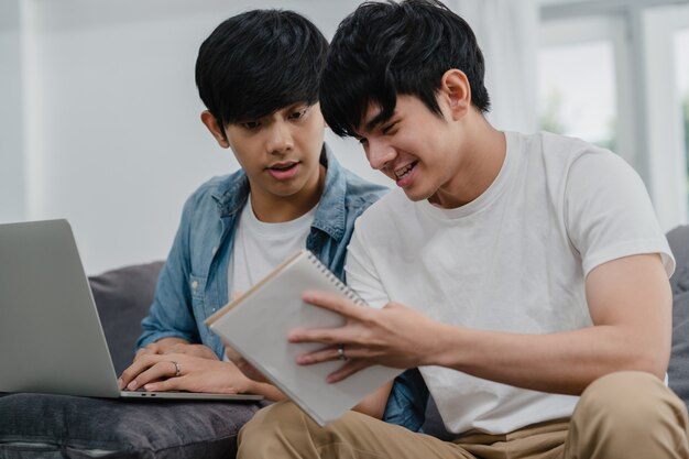 Young Asian Gay couple working laptop at modern home. Asia LGBTQ+ men happy relax fun using computer and analyzing their finances in internet together while lying sofa in living room at house .