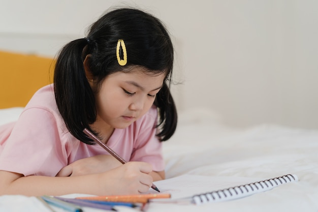 Young Asian girl drawing at home. Asia japanese woman child kid relax rest fun happy draw cartoon in sketchbook before sleep lying on bed, feel comfort and calm in bedroom at night concept.