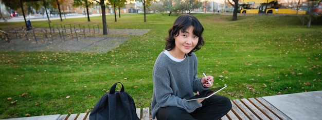 Free photo young asian girl with graphic pencil and tablet sits in park on bench draws scatches does her