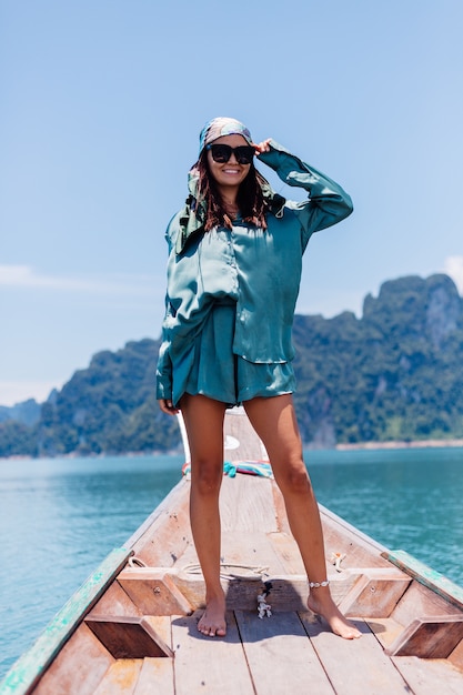 Young asian happy woman blogger tourist in silk suit and scarf and sunglasses on vacation travel around thailand on asian boat, Khao Sok national park.