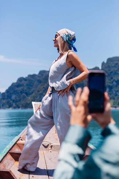 Young asian happy woman blogger tourist in silk suit and scarf and sunglasses on vacation travel around thailand on asian boat, Khao Sok national park.