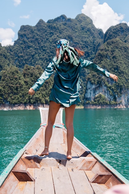 Young asian happy woman blogger tourist in silk suit and scarf and sunglasses on vacation travel around thailand on asian boat, Khao Sok national park.