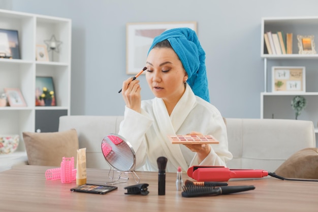 Free photo young asian happy woman with towel on her head sitting at the dressing table at home interior applying eyeshadows looking at mirror doing morning makeup routine beauty and facial cosmetics concept