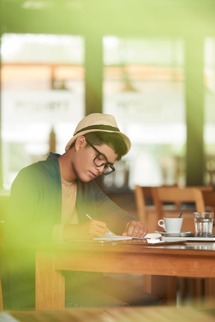 Young Asian hipster sitting in cafe and writing in notebook