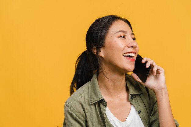 Young Asian lady talk by phone with positive expression, smile broadly, dressed in casual clothing feeling happiness and stand isolated on yellow wall. Happy adorable glad woman rejoices success.