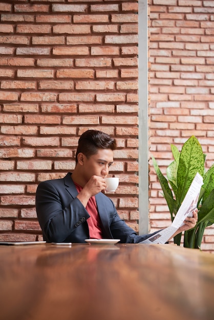 Free photo young asian man sitting at table in loft cafe, drinking coffee and reading newspaper