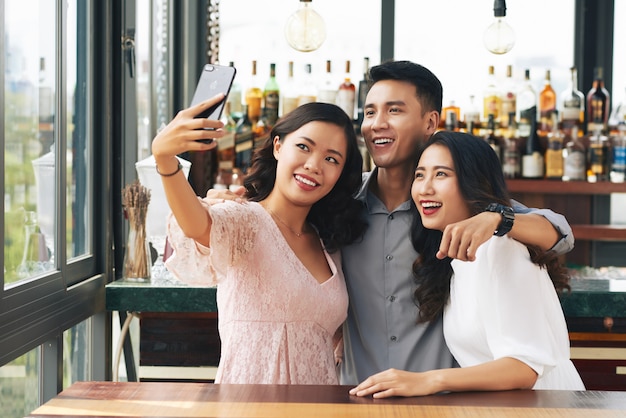 Young Asian man and two women hugging and taking selfie on smartphone in bar