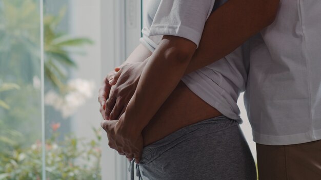 Young Asian Pregnant couple hug and holding belly talking with their child. Mom and Dad feeling happy smiling peaceful while take care baby, pregnancy near window in living room at home .