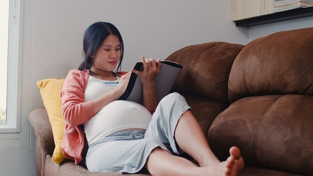 Young Asian Pregnant woman drawing baby in belly in notebook. Mom feeling happy smiling positive and peaceful while take care child lying on sofa in living room at home .