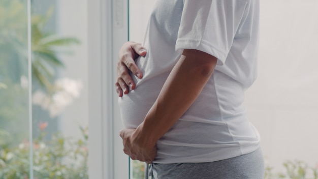 Young Asian Pregnant woman holding her belly talking with her child. Mom feeling happy smiling positive and peaceful while take care baby, pregnancy near window in living room at home .