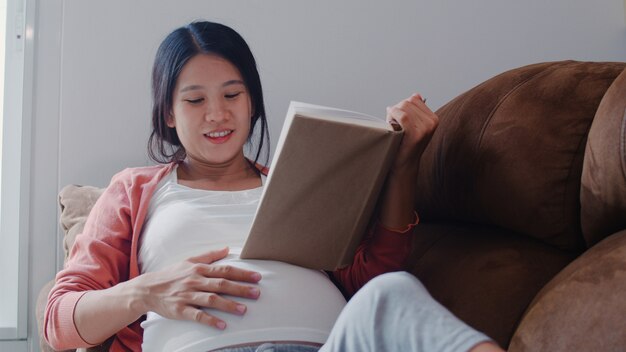 Young Asian Pregnant woman read a book for baby in belly. Mom feeling happy smiling positive and peaceful while take care child lying on sofa in living room at home .