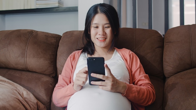 Young Asian Pregnant woman using mobile phone search pregnancy information. Mom feeling happy smiling positive and peaceful while take care her child lying on sofa in living room at home .