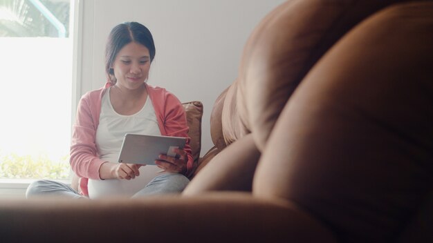 Young Asian Pregnant woman using tablet search pregnancy information. Mom feeling happy smiling positive and peaceful while take care her child lying on sofa in living room at home .